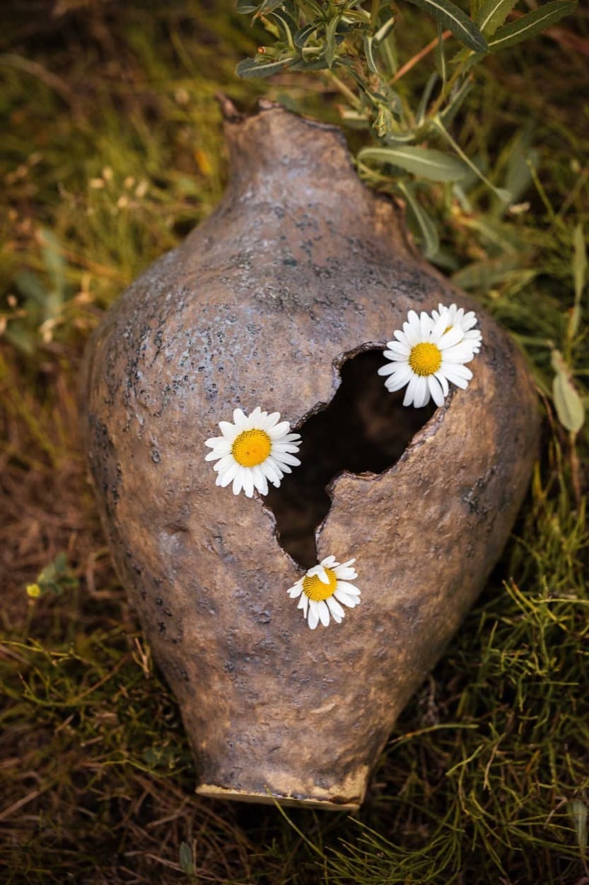 Alex Roby ceramic vessel with a broken opening and daisies, lying in the grass