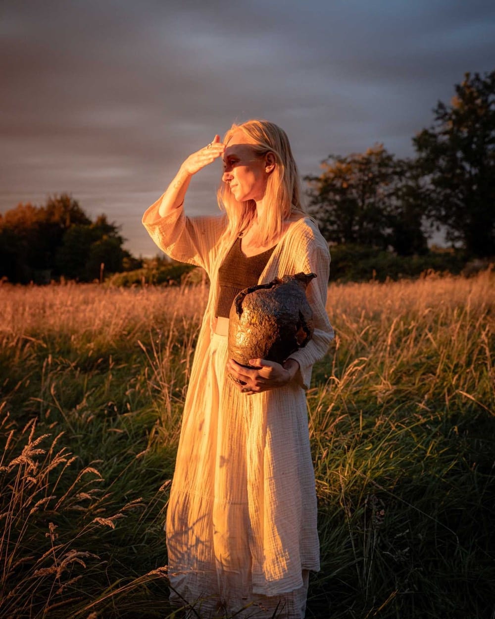 Alex Roby shading her eyes in a field at sunset, a dark vessel in her arm