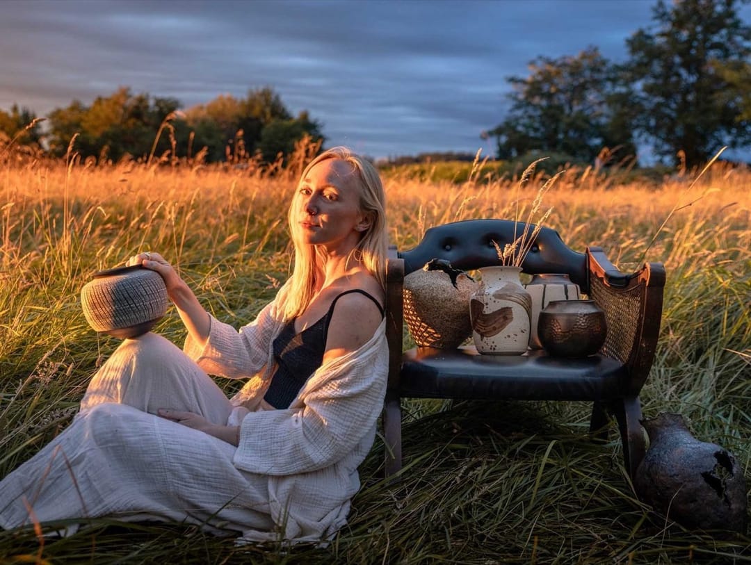 Alex Roby in a golden field at sunset, holding a bowl beside a bench of her pottery