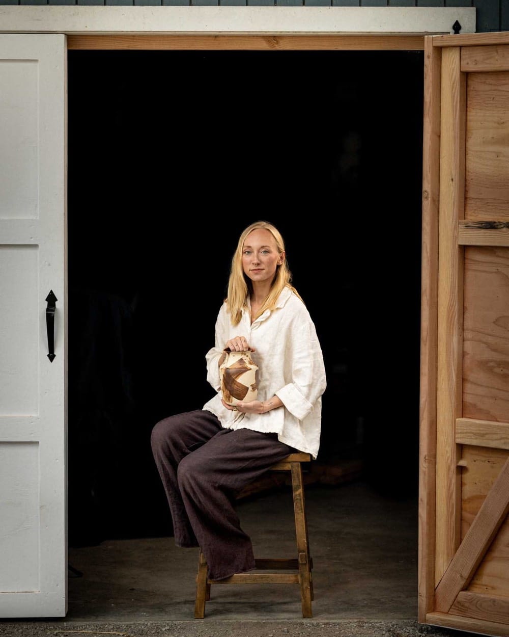 Alex Roby on a stool in her studio doorway, holding a ceramic piece