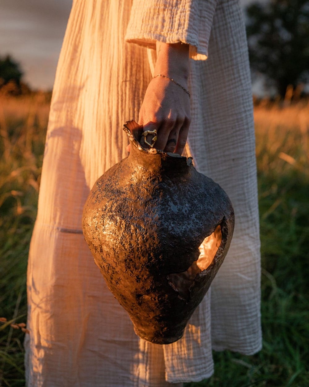 Hand holding a dark metallic ceramic vessel against a white linen dress at golden hour