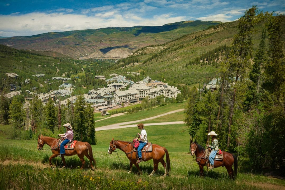 Tres jinetes a caballo en una pradera verde sobre el valle, con Park Hyatt Beaver Creek a lo lejos