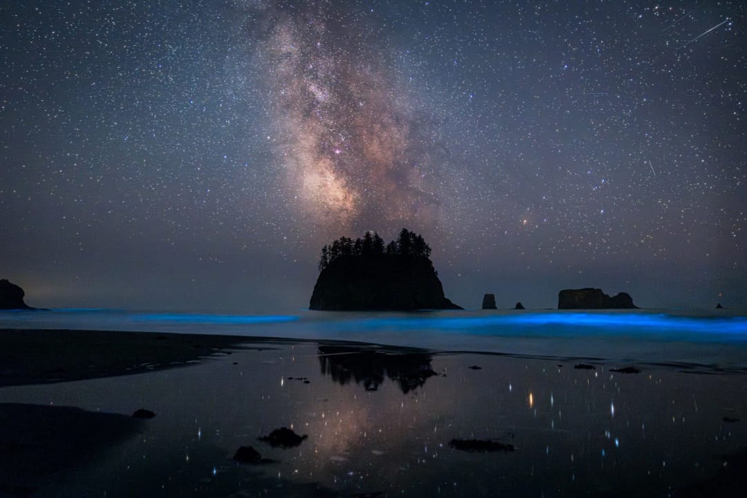 Cosmic Beach. La Vía Láctea sobre un farallón, olas que brillan en azul y su reflejo en la arena mojada