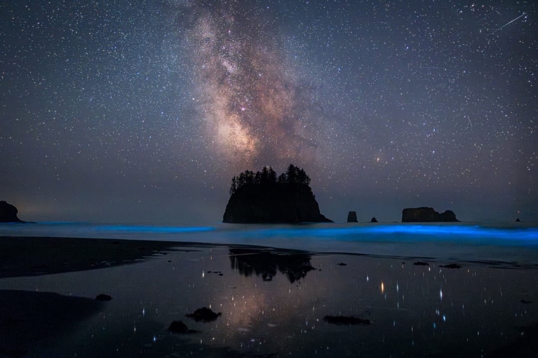 Cosmic Beach. La Vía Láctea sobre un farallón, olas que brillan en azul y su reflejo en la arena mojada