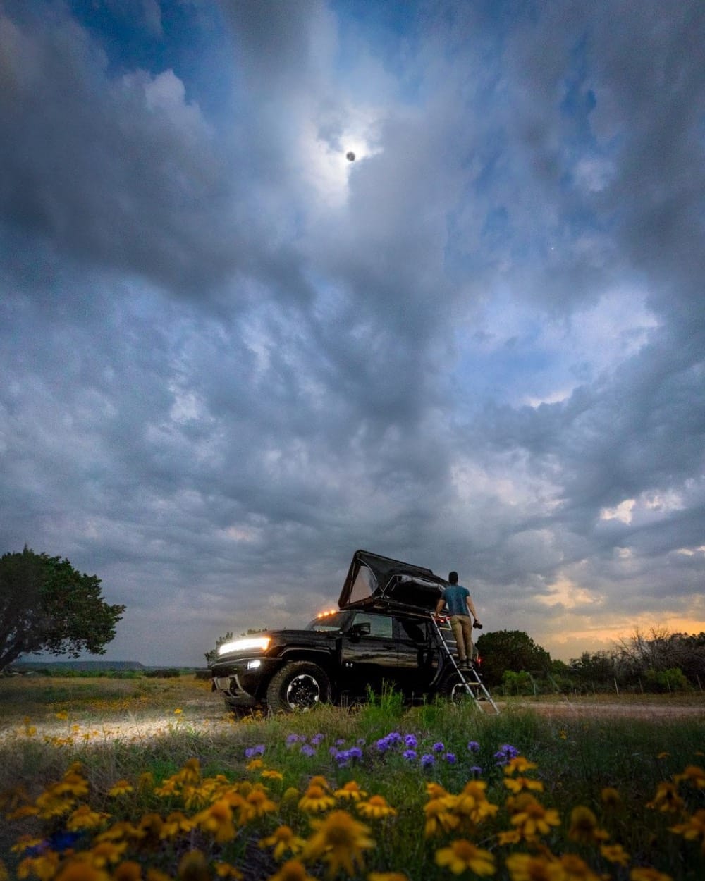GMC Hummer EV con carpa de techo bajo el eclipse solar total de 2024 en Texas, un hombre en la escalera y flores silvestres al frente