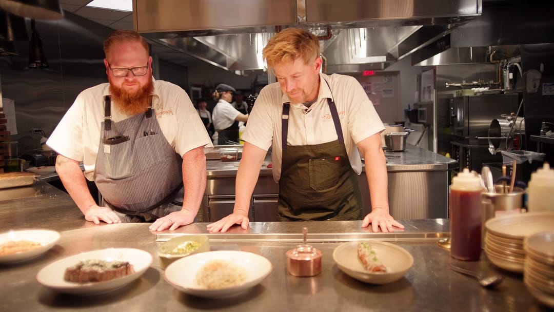 El chef Blais y otro chef en el pase de la cocina, con platos servidos al frente, del video de los restaurantes