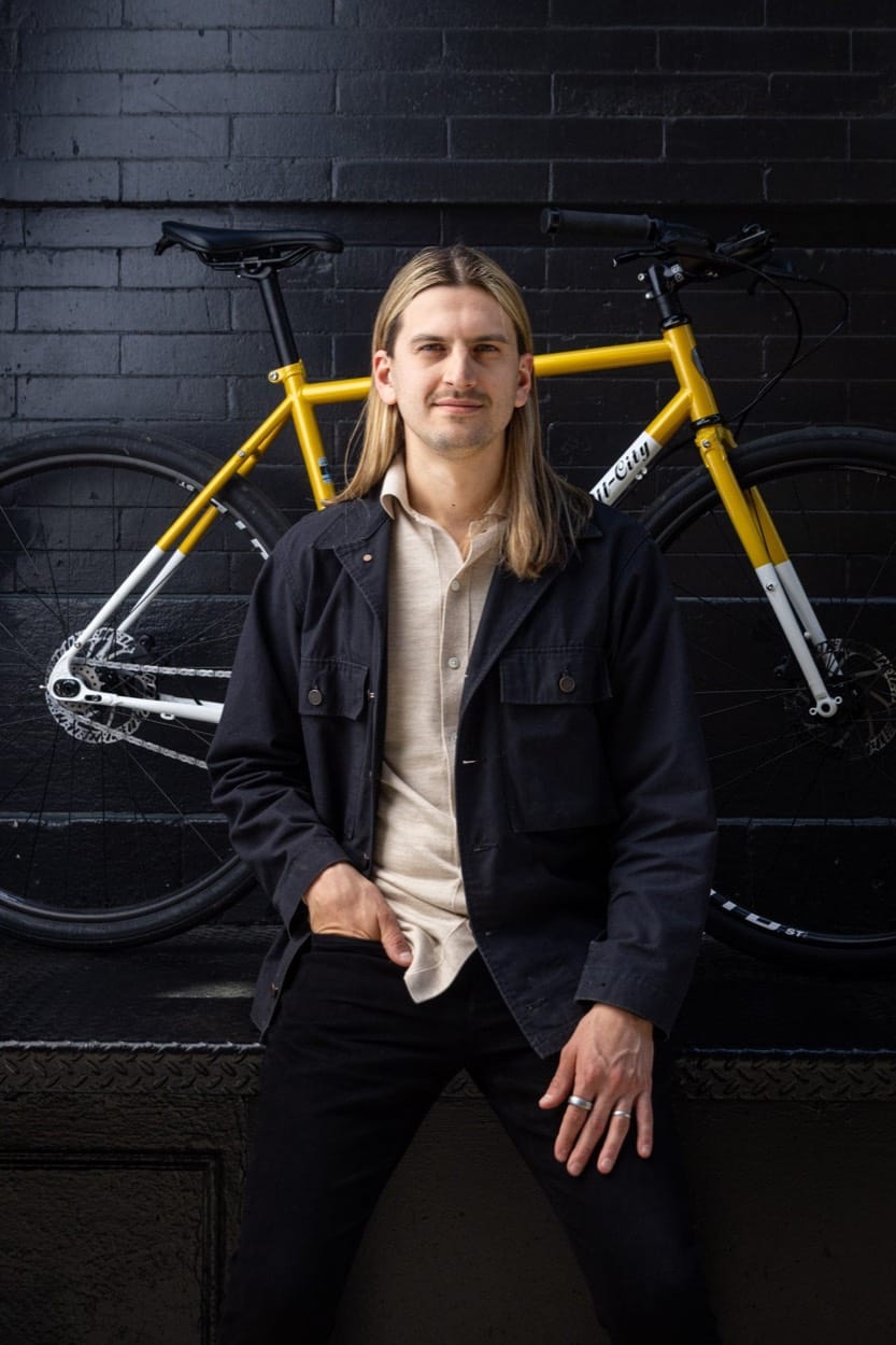 Harkov Lewis Team portrait, a man in front of a yellow bike on a black brick wall