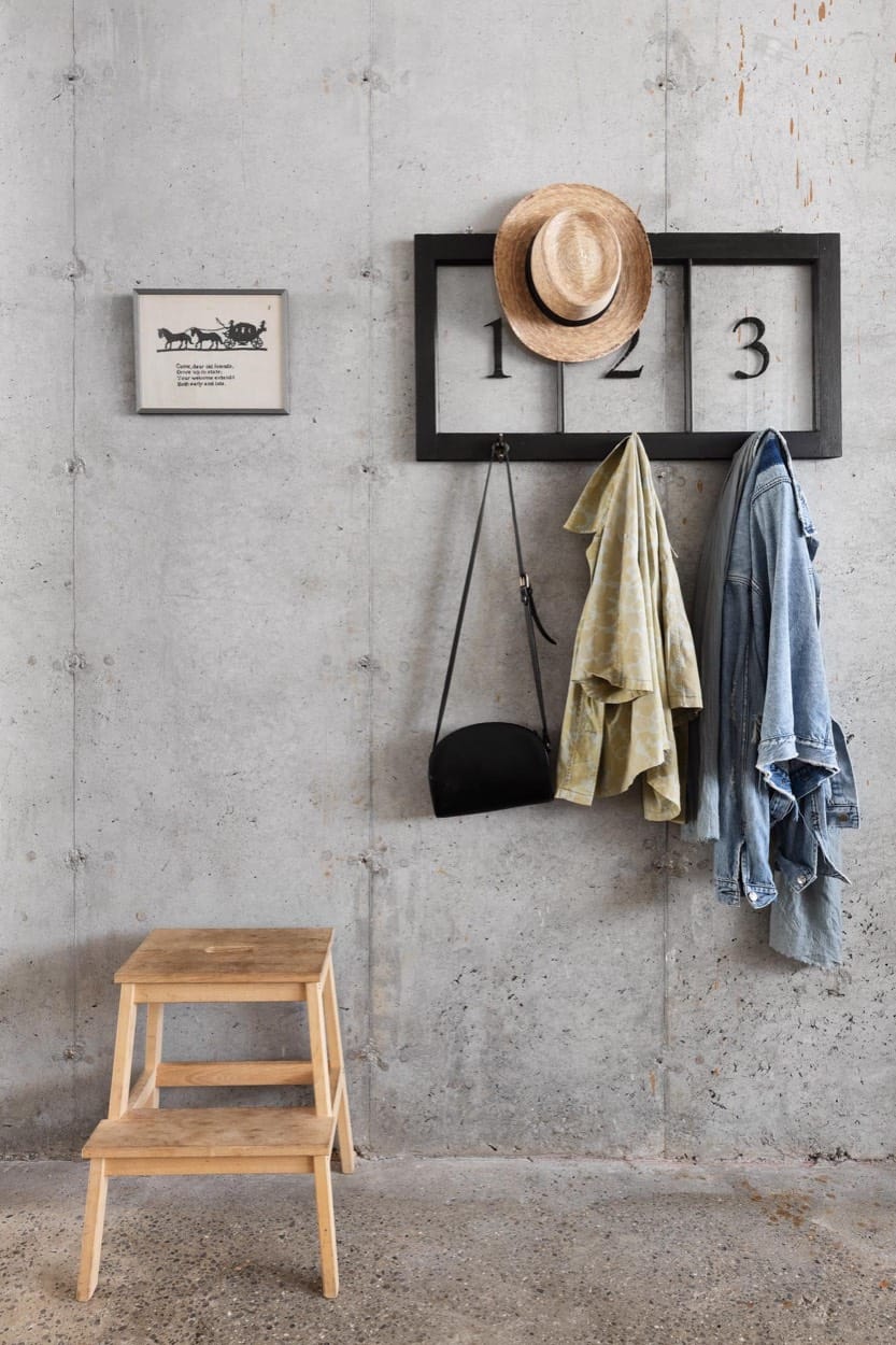Muro de concreto con ganchos numerados, un sombrero de paja, chaquetas, un bolso negro y un banquito de madera