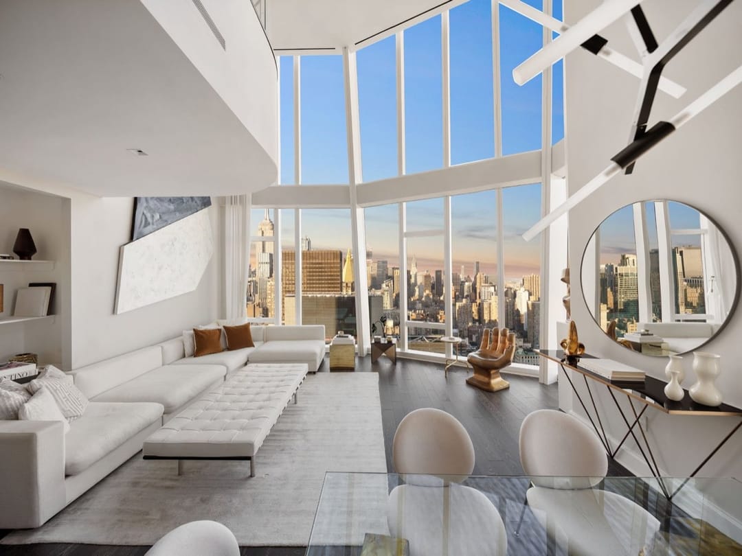 Sala de doble altura con sofás blancos y ventanales de piso a techo hacia Manhattan