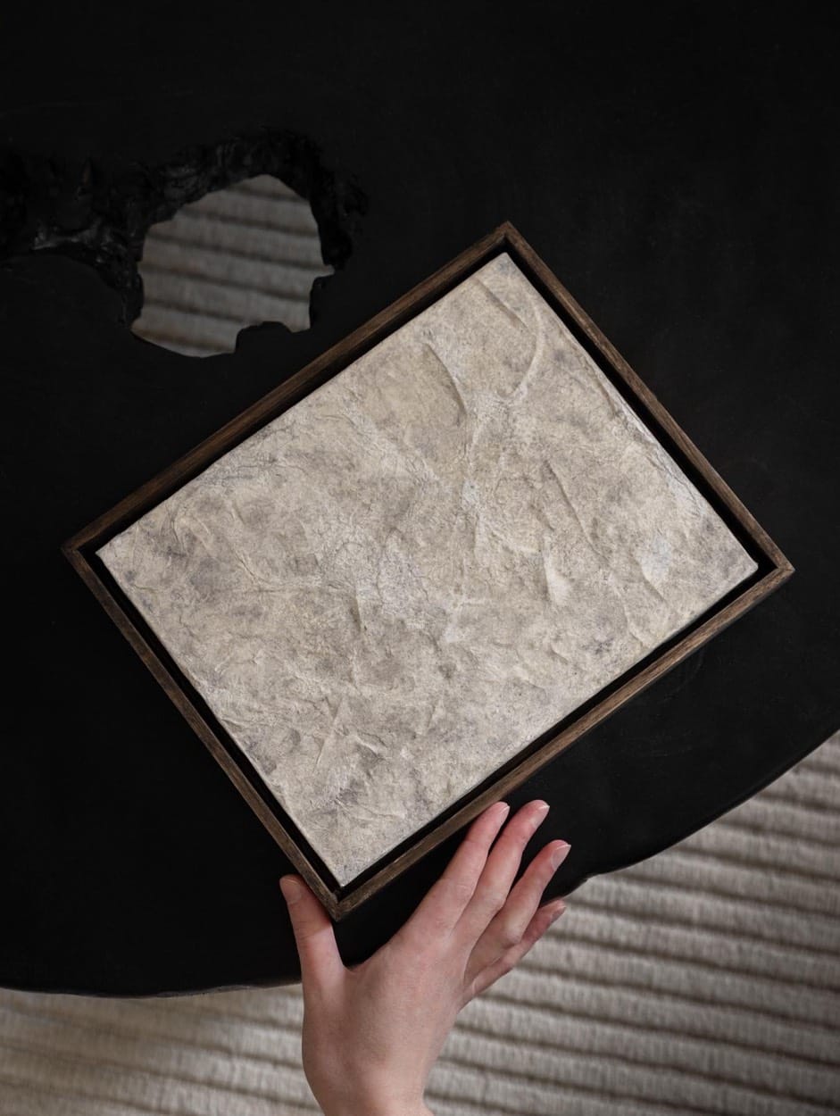 Obra enmarcada con textura de piedra de Jane Kidder sobre una mesa negra, una mano en la esquina