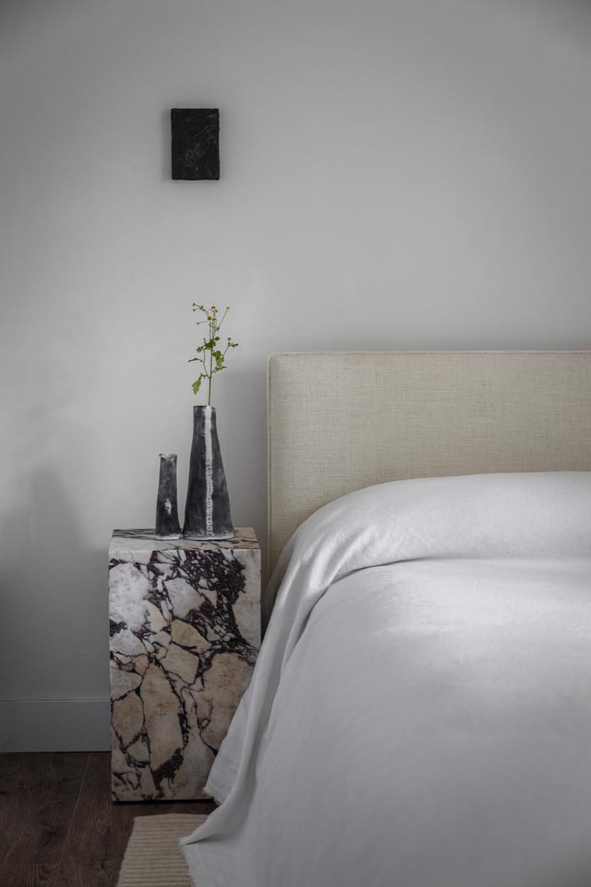Dormitorio con mesa de noche de mármol en forma de cubo, un jarrón negro y una cabecera tapizada