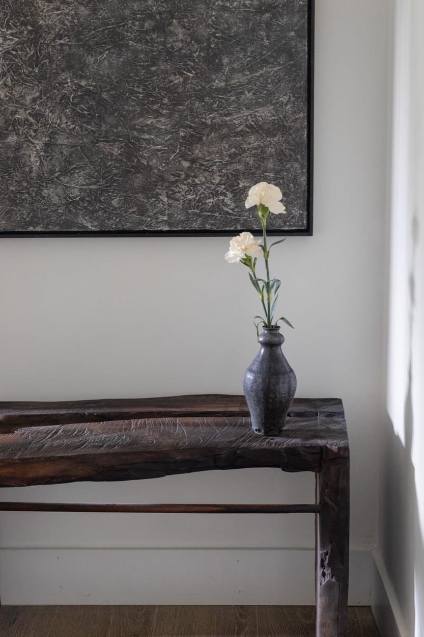 Rustic wood console with a black vase of white carnations below a dark textured artwork
