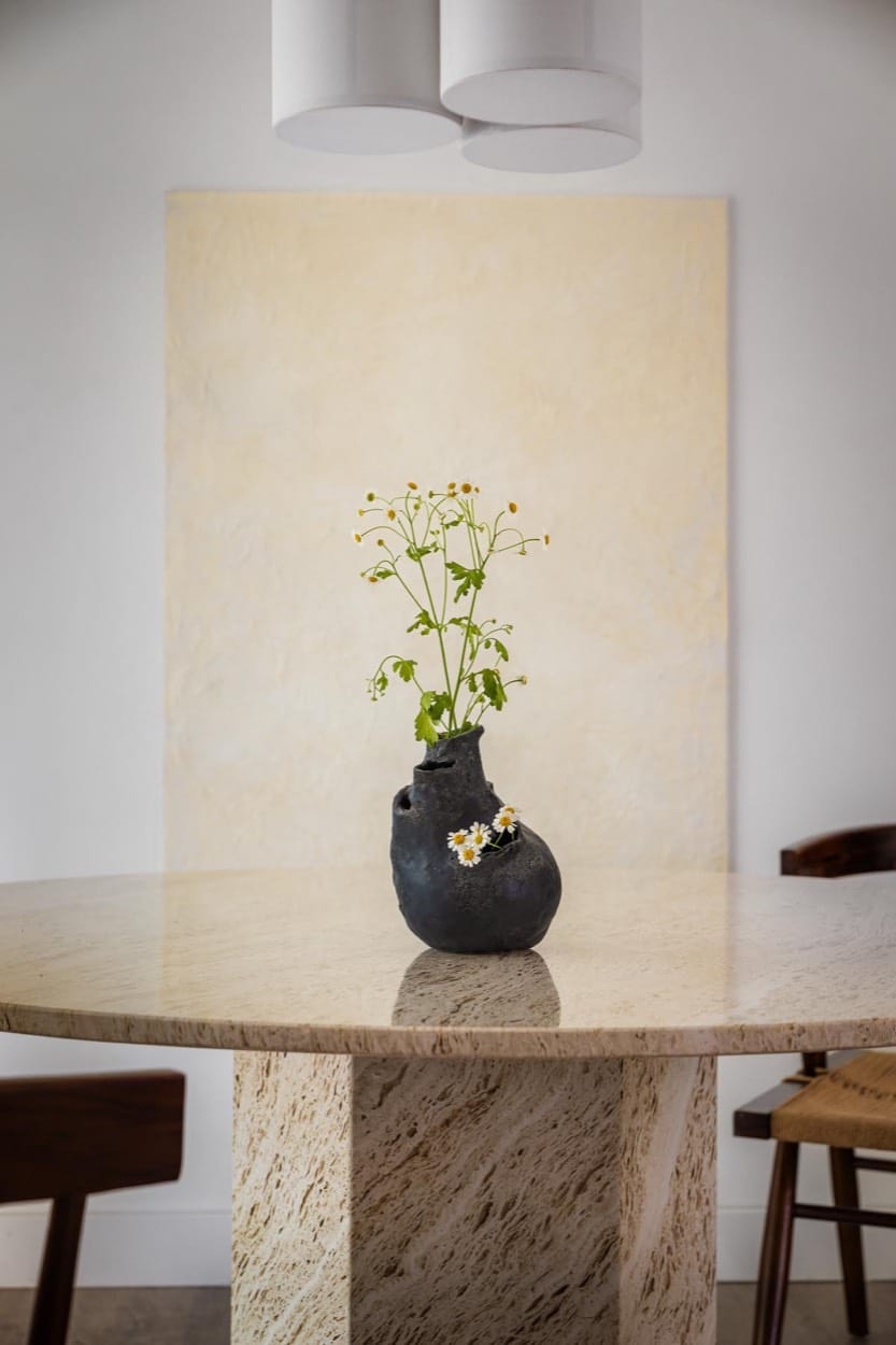 Mesa de comedor con pedestal de piedra y un jarrón negro con flores blancas, un lienzo claro detrás