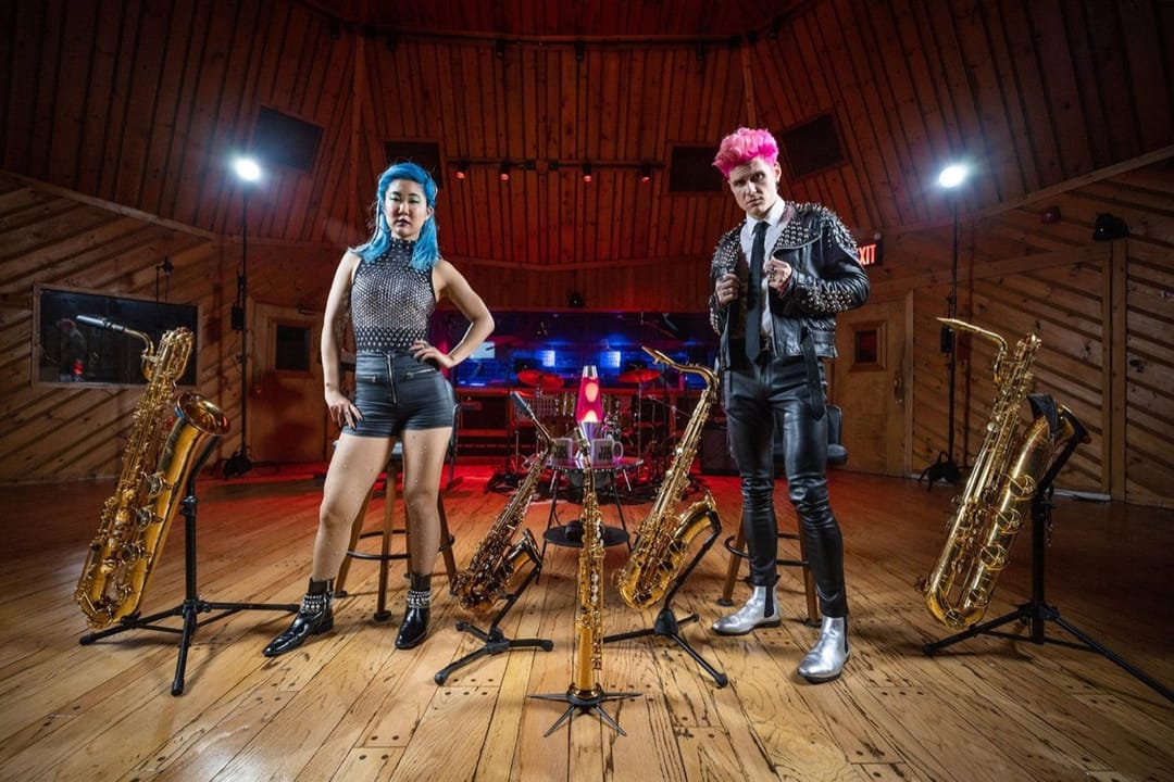 Dos músicos de cabello azul y rosa entre saxofones en un estudio de grabación con paneles de madera