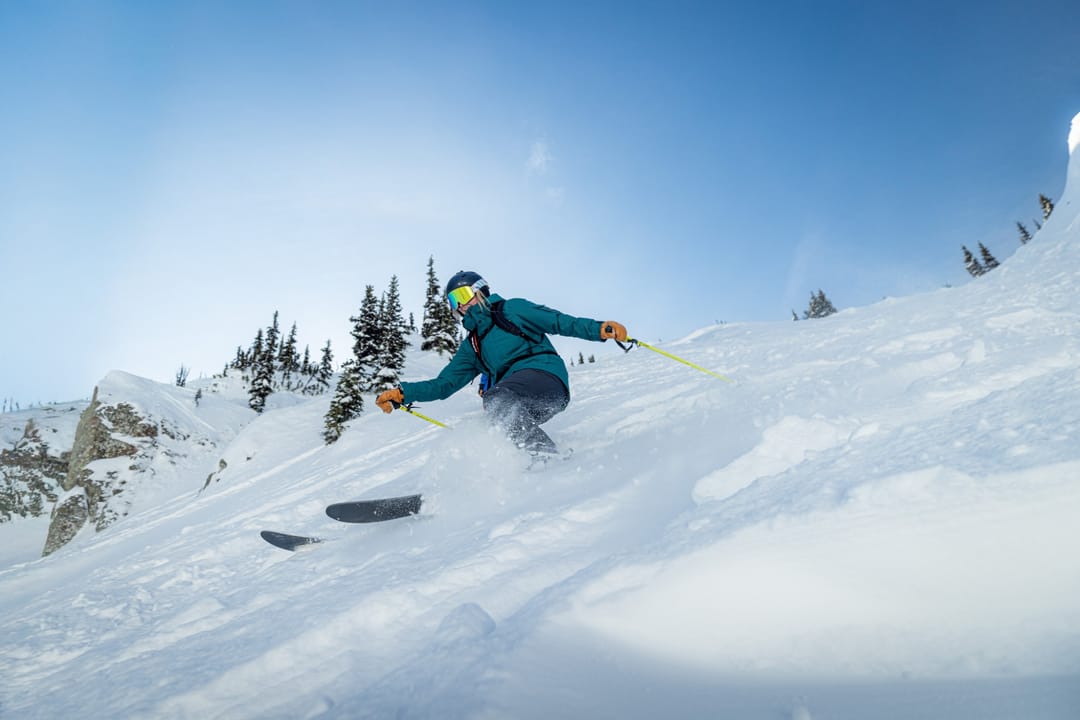 Esquiadora con chaqueta verde azulado trazando una curva en nieve polvo bajo pinos nevados