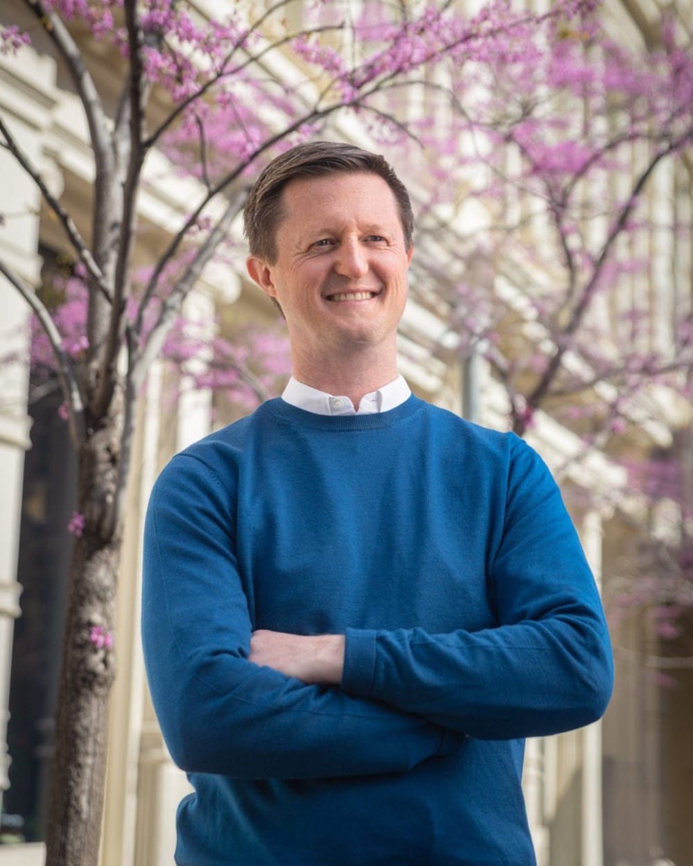 Team branding portrait in SoHo, a man in a blue sweater with arms crossed under a tree in pink bloom