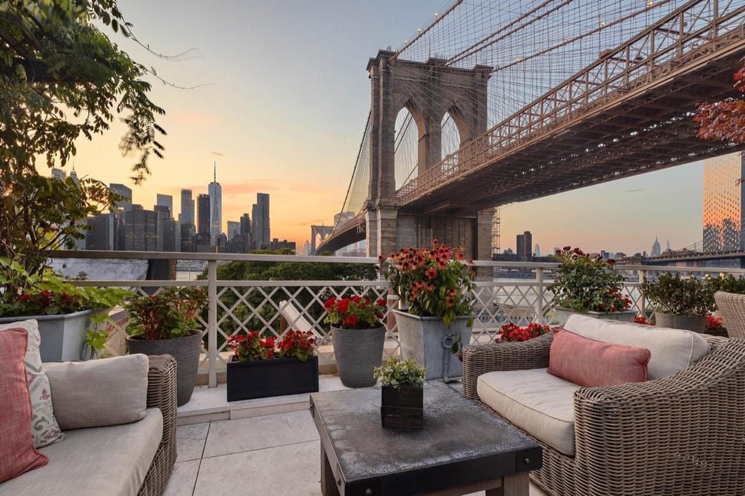 Terraza con sofás de mimbre y flores rojas bajo el puente de Brooklyn, con el skyline de Manhattan al atardecer